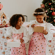 Two young girls in matching outfits standing in front of a Christmas tree with gift boxes.